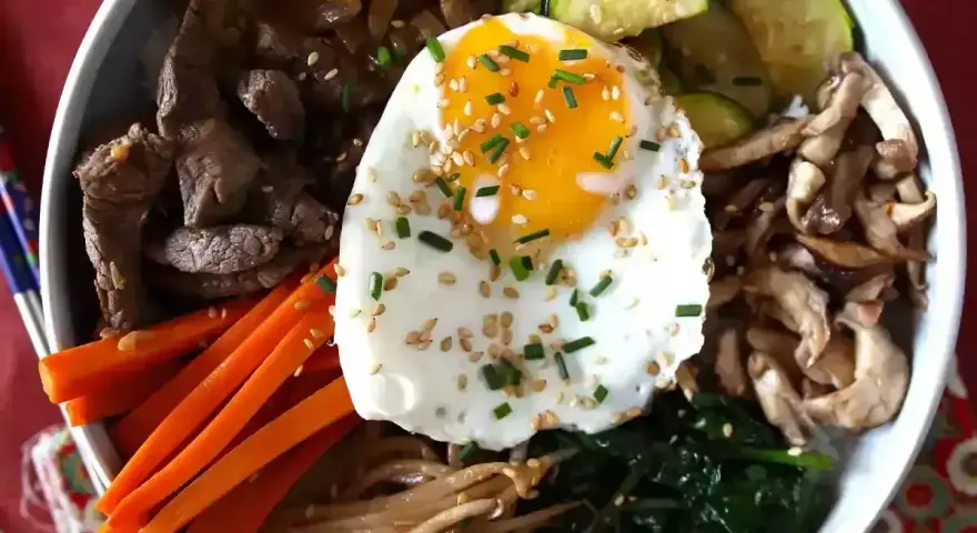 Apetyczne koreańskie dania: bibimbap z wołowiną, warzywami, grzybami i jajkiem sadzonym, posypane sezamem i szczypiorkiem.