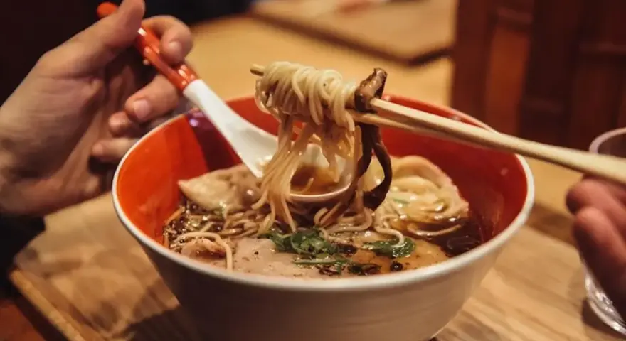 Ręka trzyma pałeczkami porcję parującego ramenu z makaronem i grzybami. Oto jak jesc ramen!