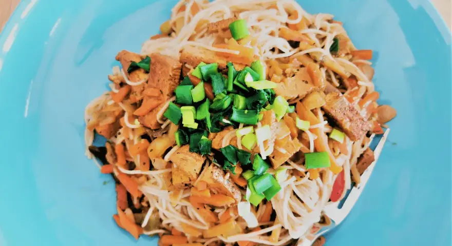 Pyszne tofu po chińsku z makaronem ryżowym, warzywami i szczypiorkiem na niebieskim talerzu.