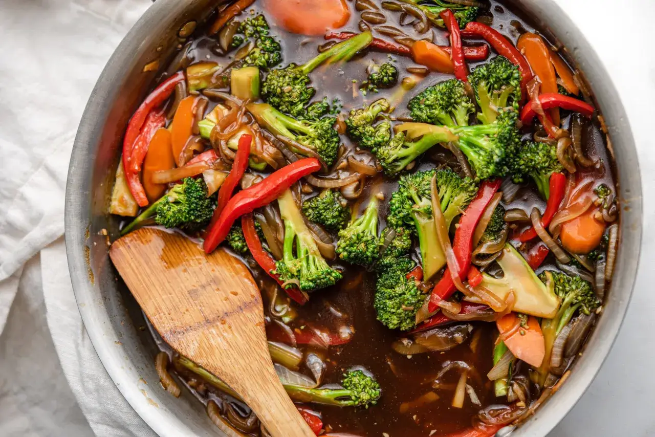 Kolorowe warzywa w sosie teriyaki: brokuły, papryka i marchewka gotujące się na patelni. Drewniana łopatka miesza danie.