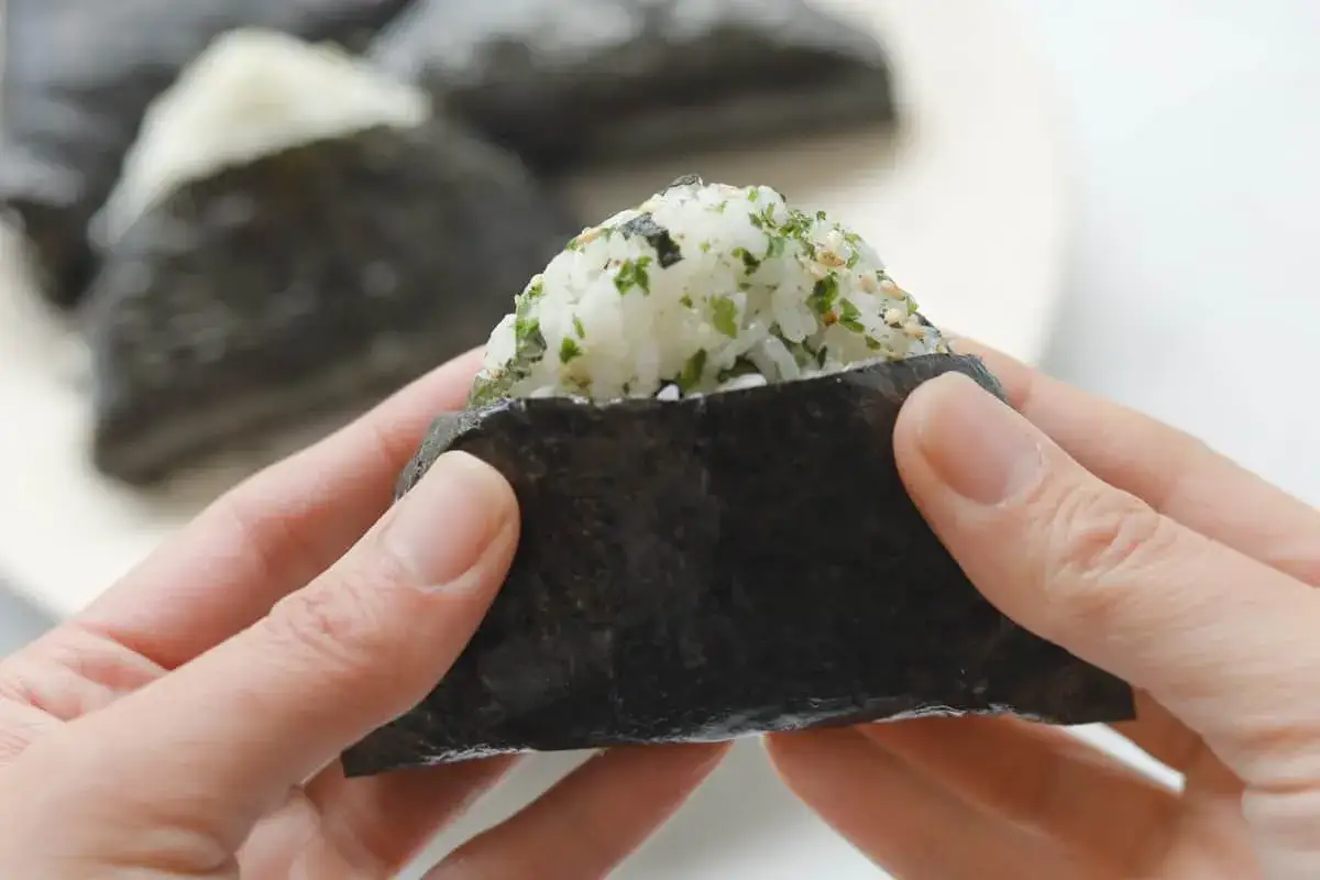 Dłonie trzymają onigiri, ryżowe trójkąty zawinięte w nori. Zastanawiasz się, którą stroną nori do sushi? Ta jest gotowa do jedzenia!
