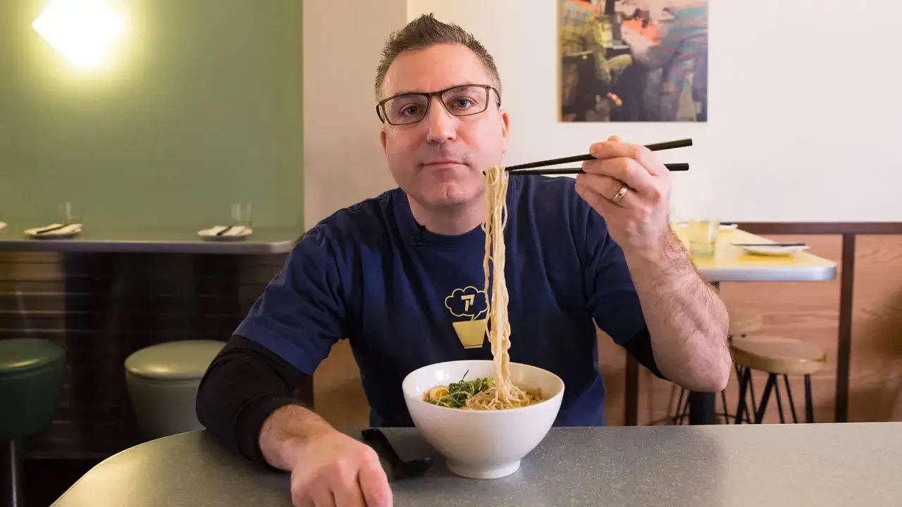 Mężczyzna z okularami pokazuje, jak jesc ramen, podnosząc pałeczkami porcję makaronu z miski.