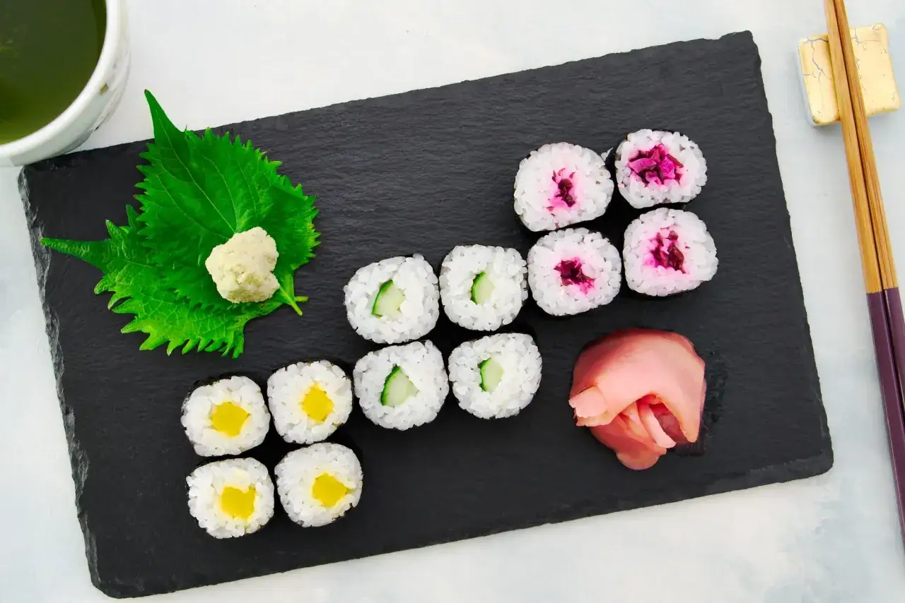 Zestaw sushi: hosomaki oshinko z żółtą rzodkiewką, ogórkiem i różowym imbirem. Obok zielona herbata i pałeczki.