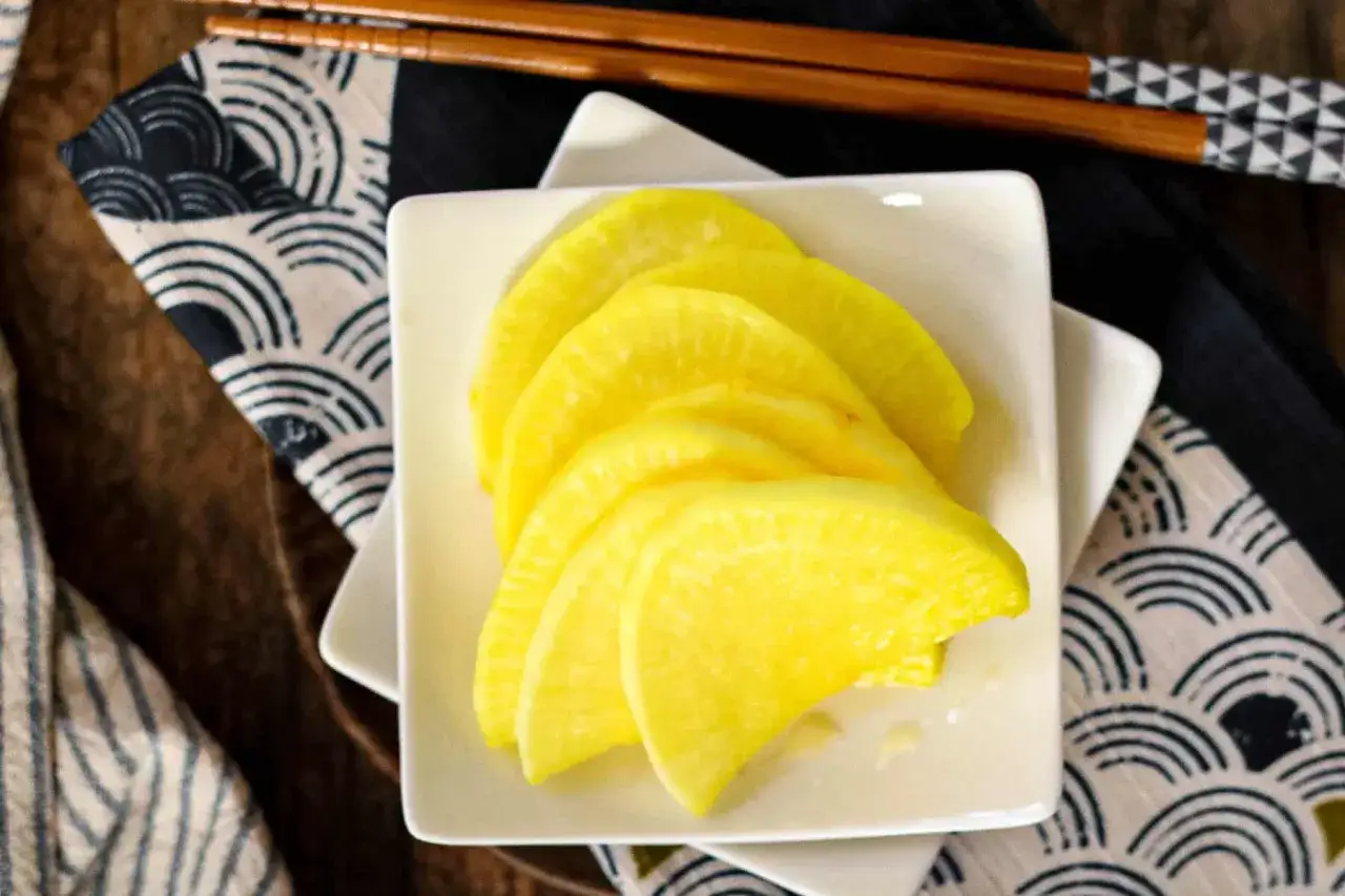 Żółta rzodkiew do sushi, pokrojona w plasterki, leży na białym kwadratowym talerzyku. Obok pałeczki i japoński wzór.