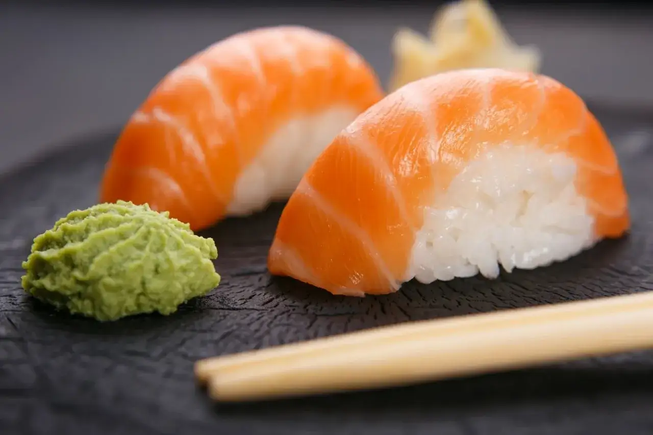 Dwa kawałki sushi z łososiem i ryżem, obok zielona pasta. Czy wasabi jest ostre? Pałeczki czekają.