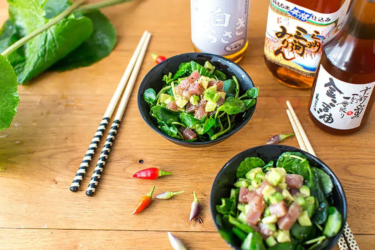 Dwa talerze z poke bowl, zielenią, awokado i tuńczykiem, obok pałeczki i butelki sosu mirin.