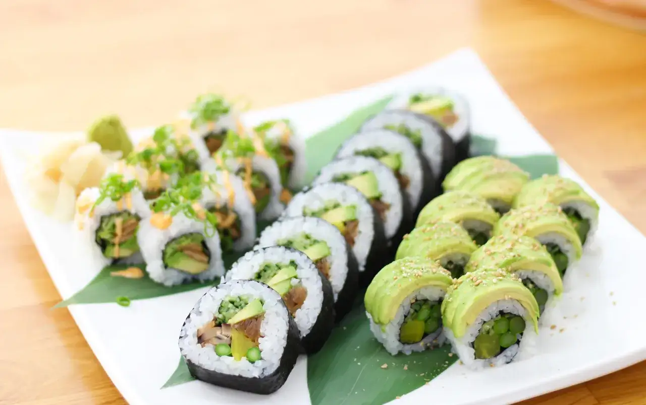 Talerz pełen kolorowych roladek sushi z warzywami: awokado, ogórek, szparagi. Obok zielona pasta wasabi i marynowany imbir.