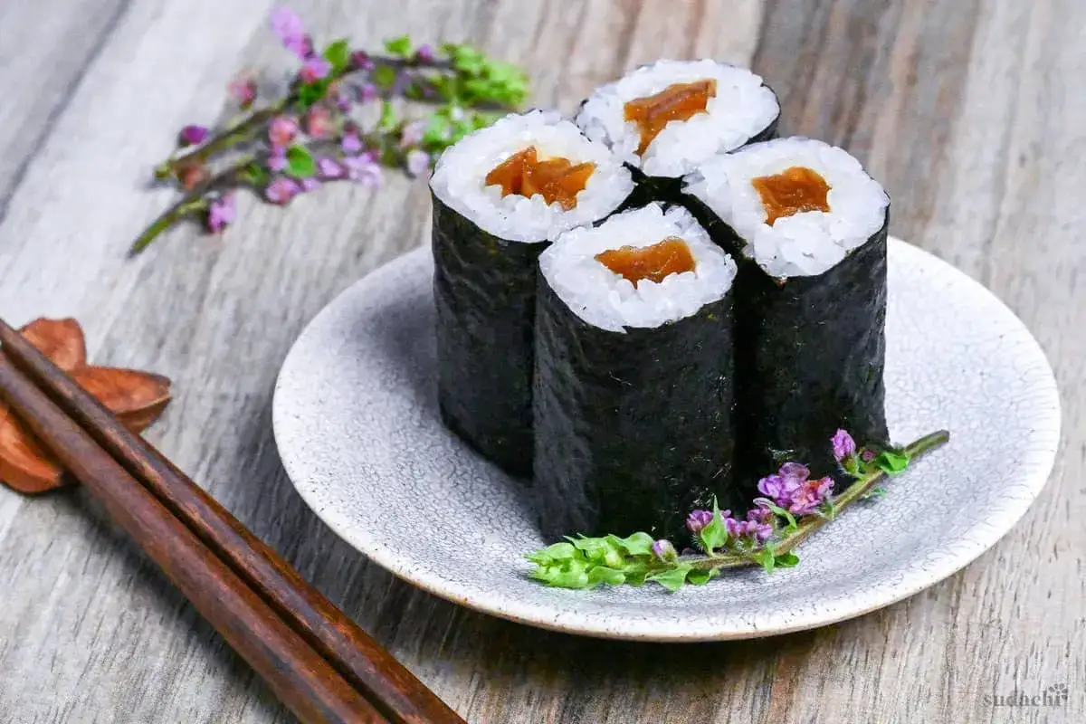 Cztery rolki kanpyo maki na talerzyku, ozdobione gałązkami z fioletowymi kwiatkami. Obok pałeczki.