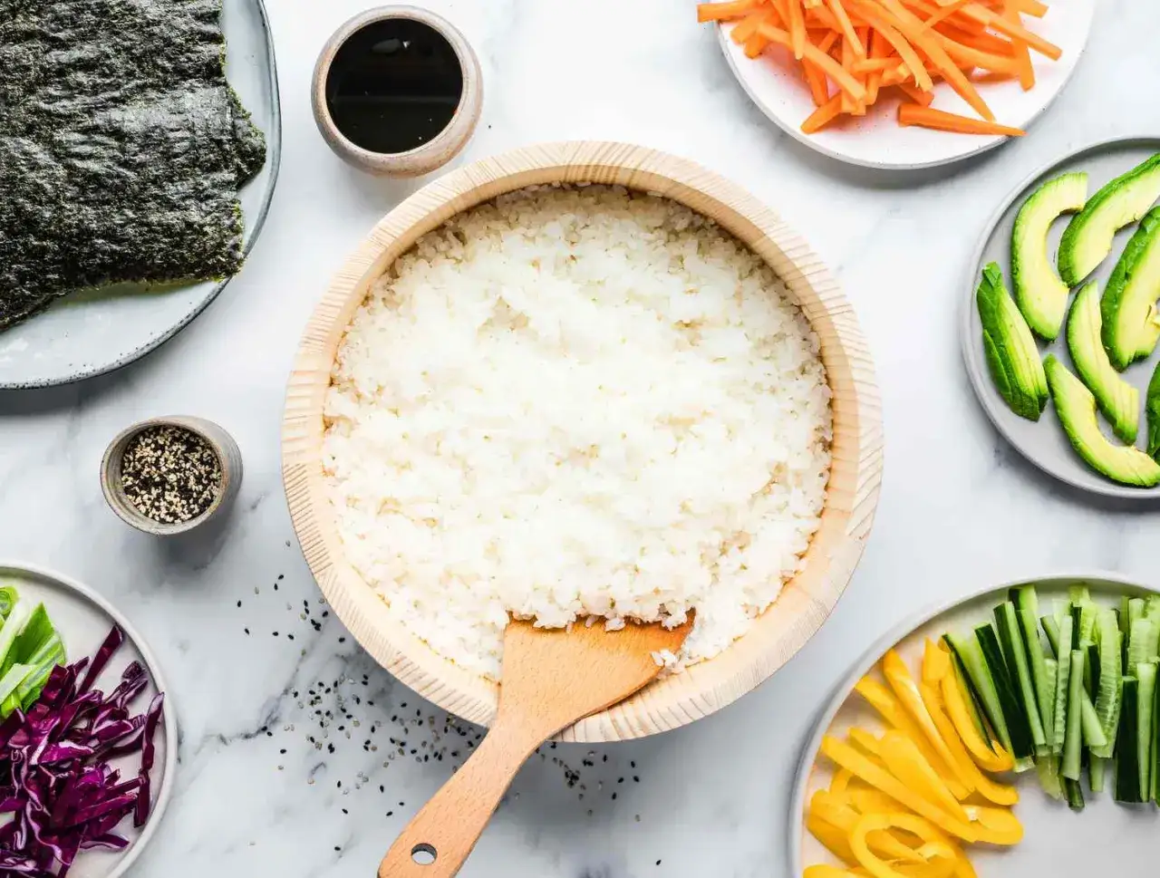 Przygotowanie ryżu do sushi: biały ryż w drewnianej misce, obok nori, sosu sojowego, sezamu, awokado, marchewki, ogórka, papryki i czerwonej kapusty. Czy ryż do sushi się soli?