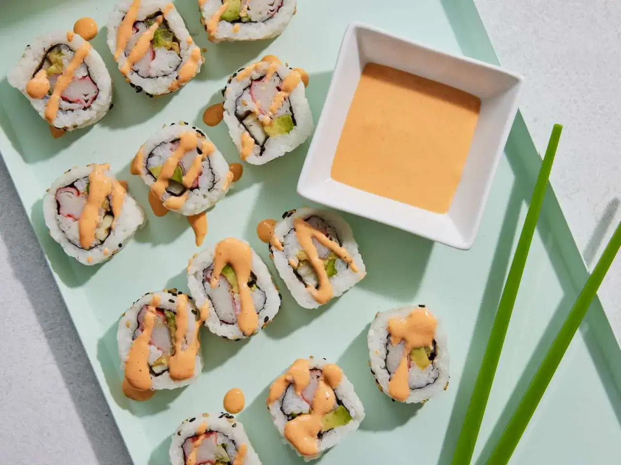 Roladki sushi z awokado i krabem, polane kremowym majonezem do sushi. Obok miseczka z sosem i pałeczki.