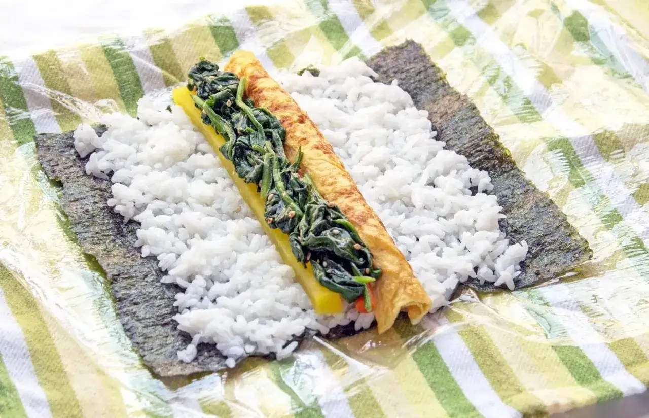 Na nori rozłożony jest ryż, a na nim żółty omlet i szpinak. Zastanawiasz się, ile ryżu na rolkę sushi?