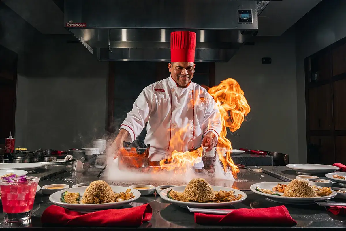 Szef kuchni w czerwonej czapce przygotowuje dania teppanyaki, tworząc spektakularny pokaz ognia i dymu.