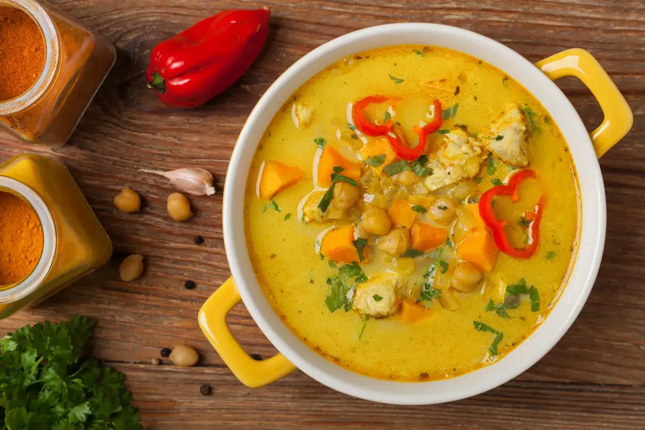Aromatyczna zupa curry z kurczakiem, ciecierzycą i dynią, ozdobiona papryczką chili. Obok słoiczki z przyprawami i świeża natka.