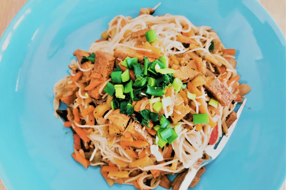 Pyszne tofu po chińsku z makaronem ryżowym, warzywami i szczypiorkiem na niebieskim talerzu.