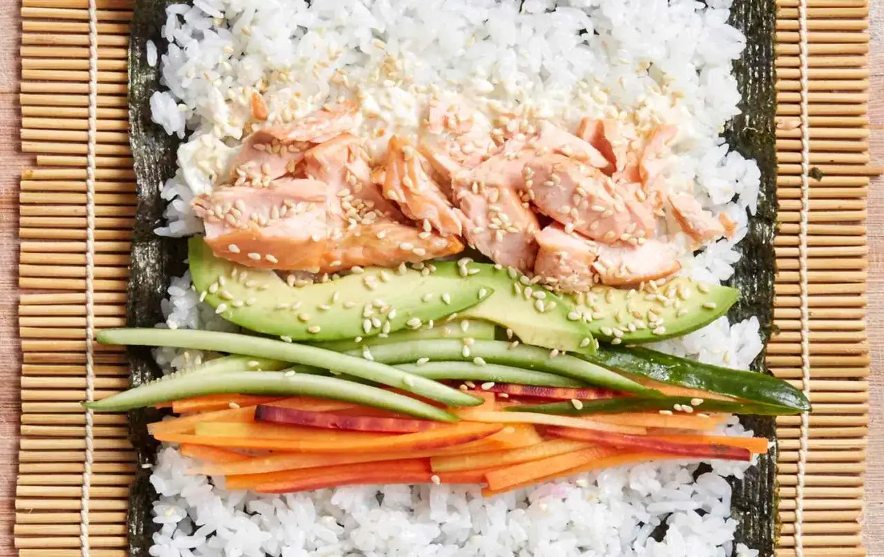 Rolka sushi z łososiem w przygotowaniu: ryż, nori, awokado, ogórek, marchewka i sezam.