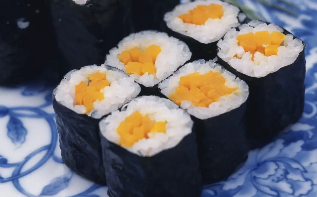Kilka kawałków sushi maki z pomarańczowym nadzieniem, prawdopodobnie oshinko sushi, ułożonych na niebiesko-białym talerzu.