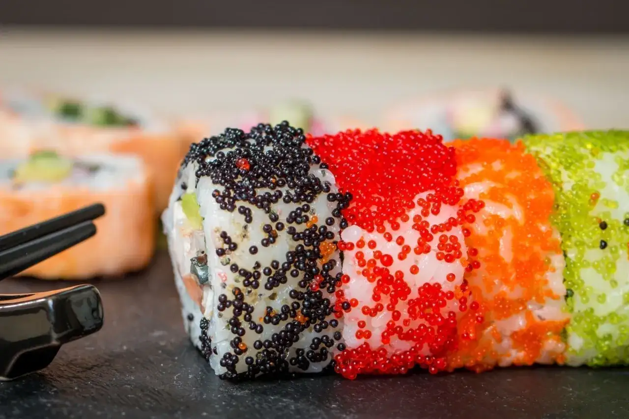 Kolorowe rolki sushi z kawiorem: czarnym, czerwonym, pomarańczowym i zielonym. Obok pałeczki i sosjerka.