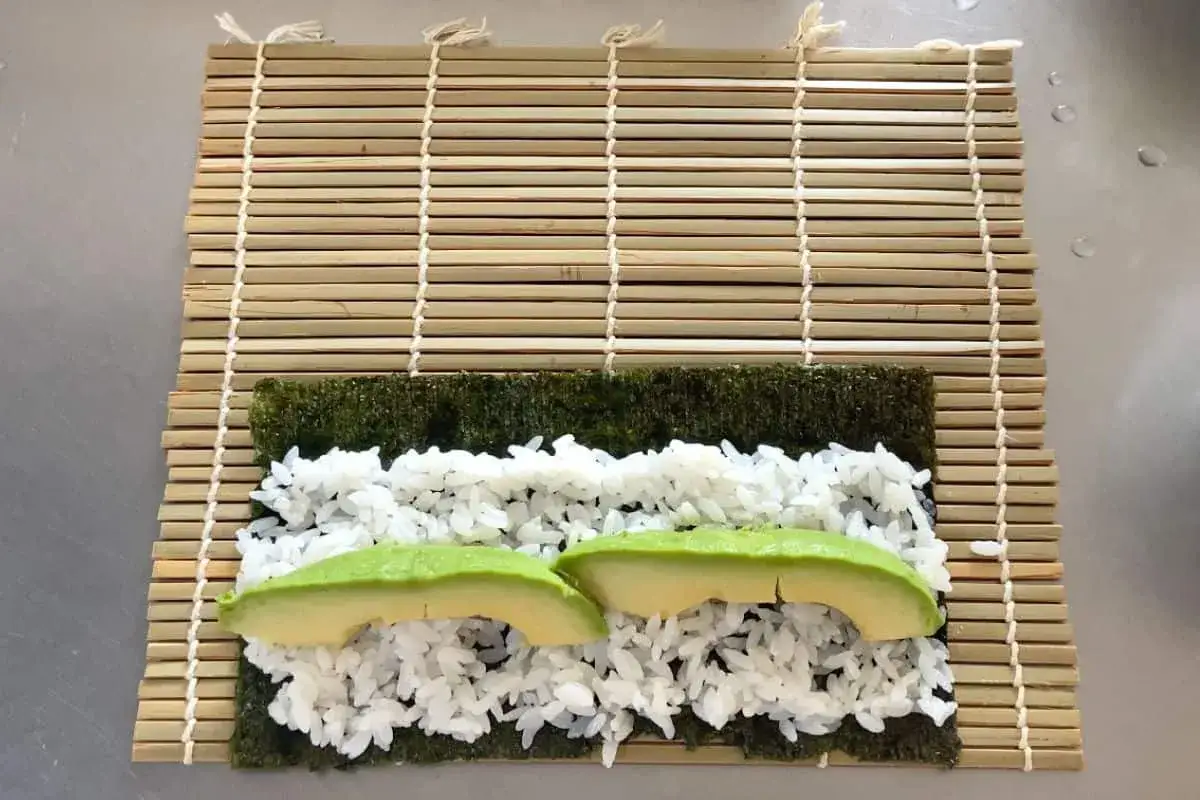 Na macie bambusowej do sushi ułożono nori, ryż i plasterki awokado do sushi.