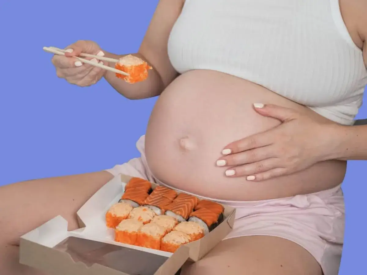 Ciężarna kobieta trzyma pałeczkami kawałek sushi, obok pudełko z rolkami.