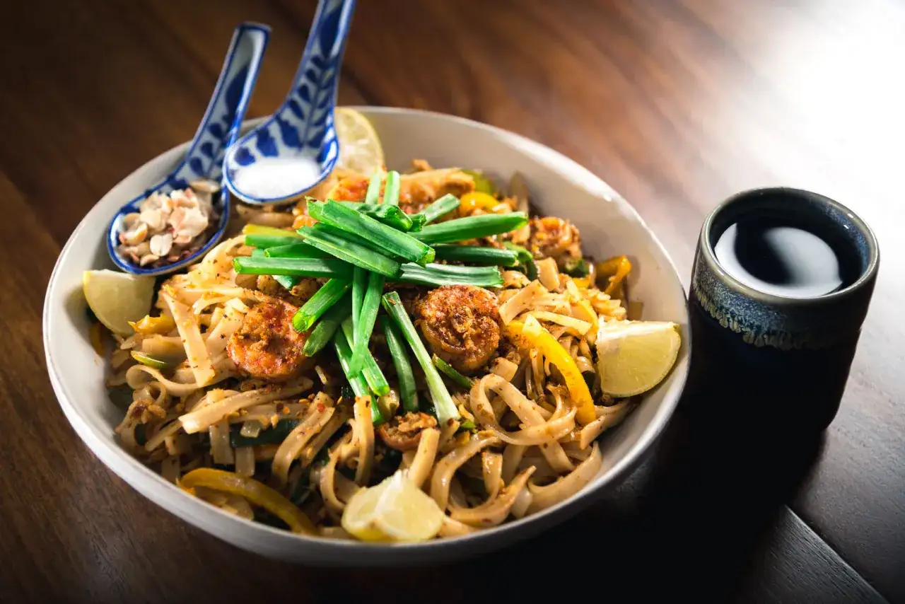 Pyszny pad thai z krewetkami, posypany szczypiorkiem i orzeszkami. Obok miseczka z sosem i cytryna. Gotowy do jedzenia!