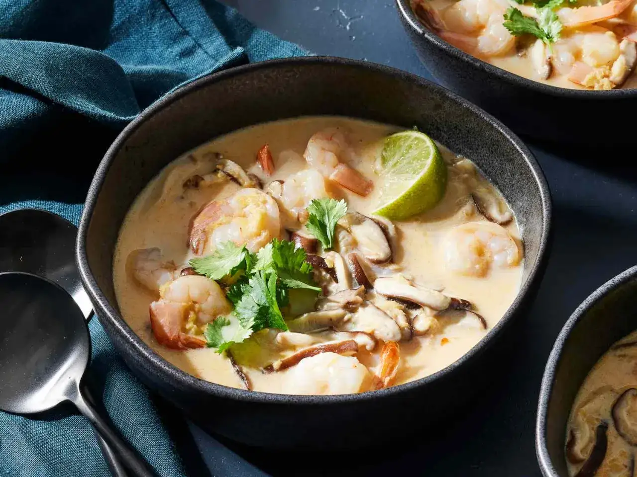 Aksamitna zupa tom kha z kurczakiem, krewetkami, grzybami i świeżą kolendrą, ozdobiona ćwiartką limonki.