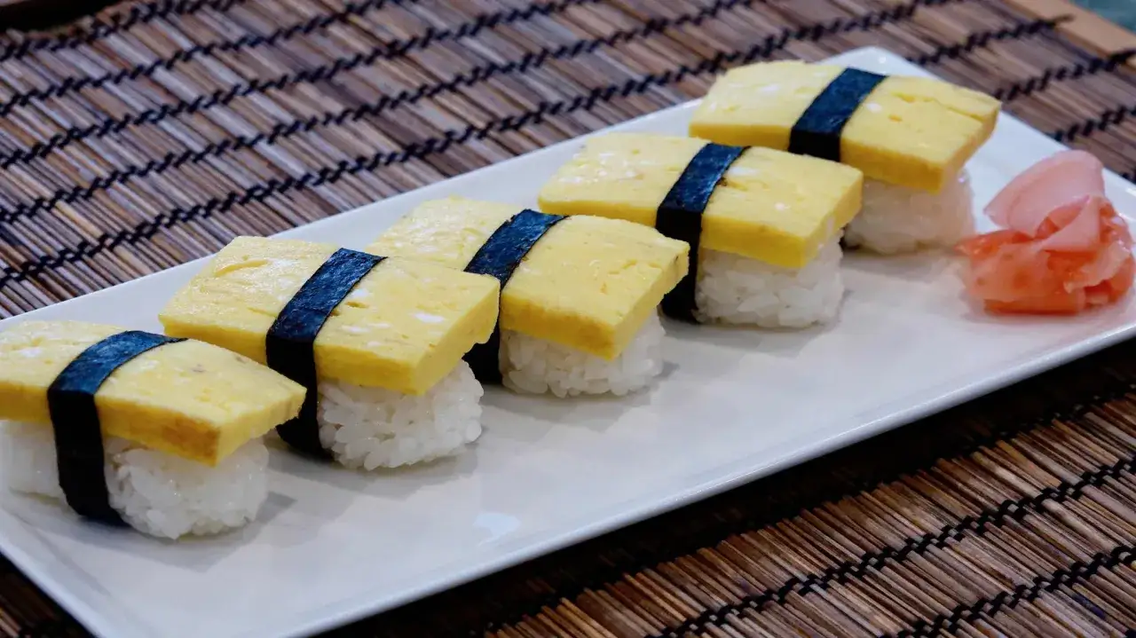 Sześć kawałków sushi datemaki z omletem na ryżu, przewiązane czarną algą nori, na białym talerzu. Obok różowy imbir.