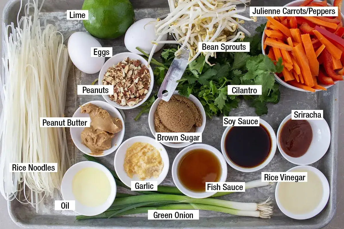 Wszystkie niezbędne pad thai składniki: makaron ryżowy, jajka, orzeszki ziemne, masło orzechowe, fasolka mung, marchewka, papryka, kolendra, sos sojowy, cukier brązowy, czosnek, olej, sos rybny, ocet ryżowy, zielona cebulka i limonka.