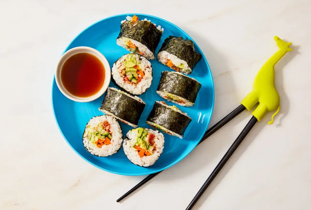 Sushi z awokado, ogórkiem i marchewką na niebieskim talerzu. Czy dzieci mogą jeść sushi? Tak, zwłaszcza takie! Pomocne pałeczki w kształcie żyrafy.