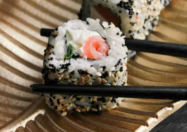 Rolka sushi z łososiem i serkiem, posypana sezamem, trzymana pałeczkami. Czy sushi można jeść na drugi dzień? Tak, jeśli było odpowiednio przechowywane.