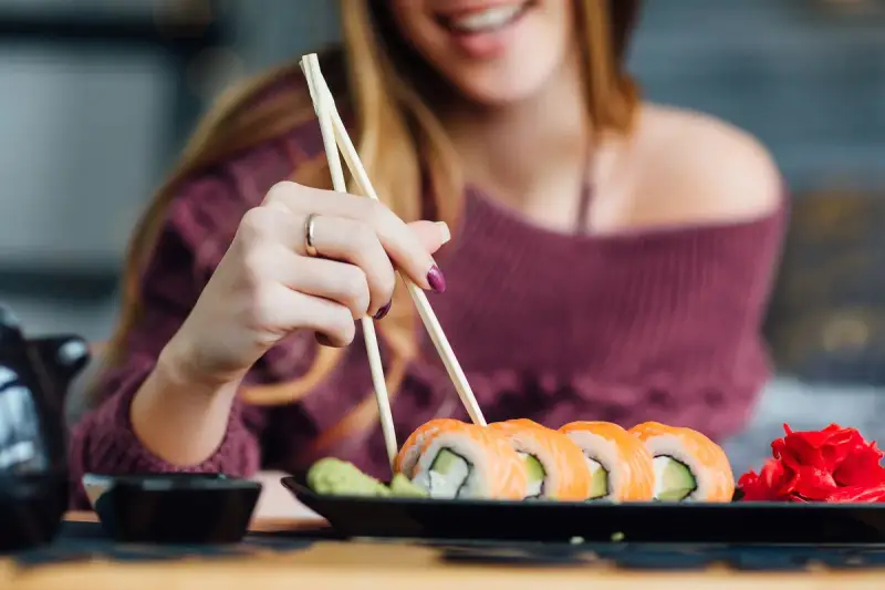 Uśmiechnięta kobieta w ciąży sięga po sushi. Na talerzu rolki z łososiem i awokado, obok wasabi i marynowany imbir.